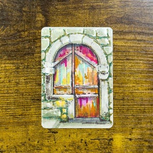 May include: A watercolor painting of a weathered wooden door with an arched stone frame. The door is painted in shades of orange, pink, and yellow, with a stone archway in shades of gray and green. The artwork is on a rectangular card.