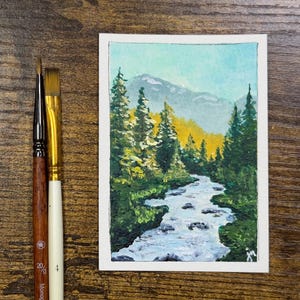 May include: A small rectangular painting of a mountain landscape featuring a flowing river, dark green pine trees, and golden foliage under a light blue sky, positioned next to two paintbrushes with wooden handles and metal ferrules.