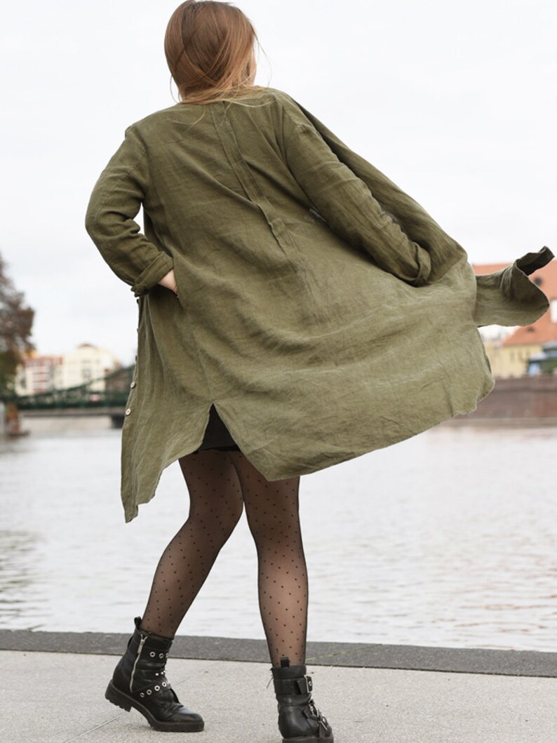 May include: A woman wearing a long, olive green linen jacket with a black polka dot patterned tights and black ankle boots.