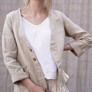 May include: A beige linen jacket with a button closure and a pocket on the left side. The jacket has a relaxed fit and three-quarter sleeves.