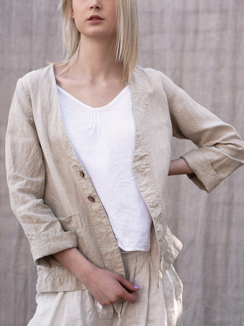May include: A beige linen jacket with a button closure and a pocket on the left side. The jacket has a relaxed fit and three-quarter sleeves.