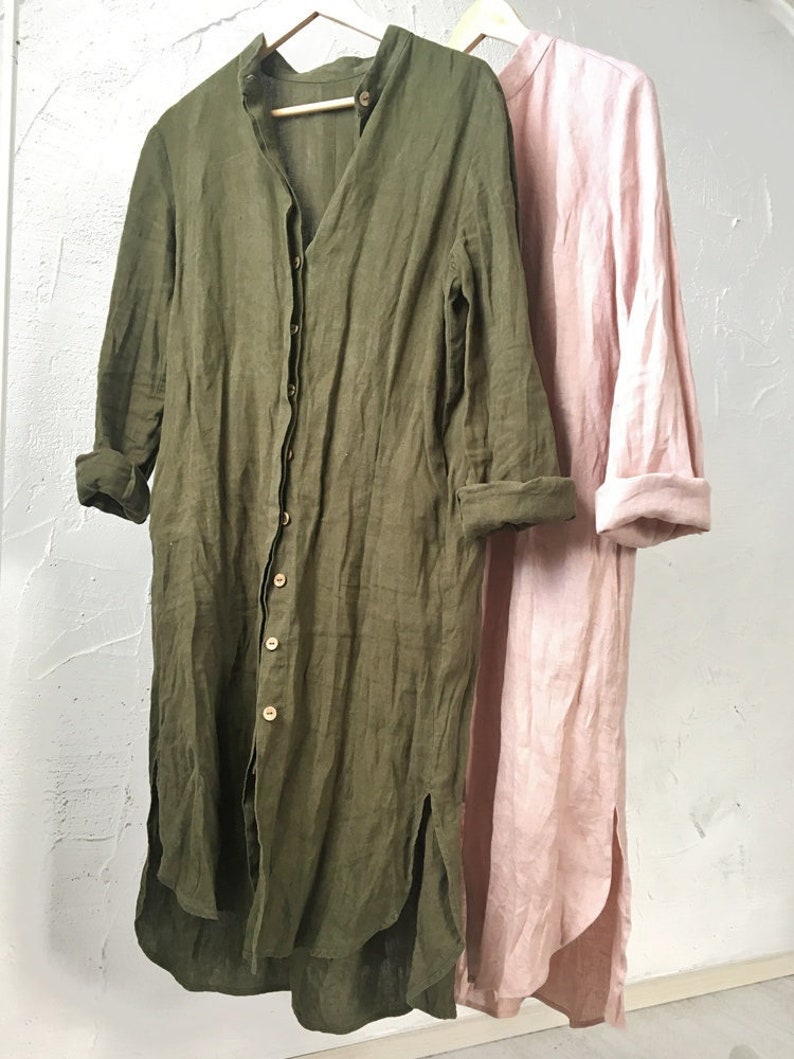 May include: Two long-sleeved linen shirts, one in a muted green and the other in a pale pink, hanging on wooden hangers against a white wall.