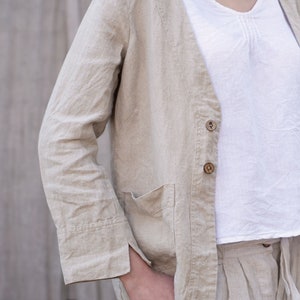 May include: A beige linen jacket with a V-neck, long sleeves, and a button closure. The jacket has a patch pocket on the left side and a single button on the front.