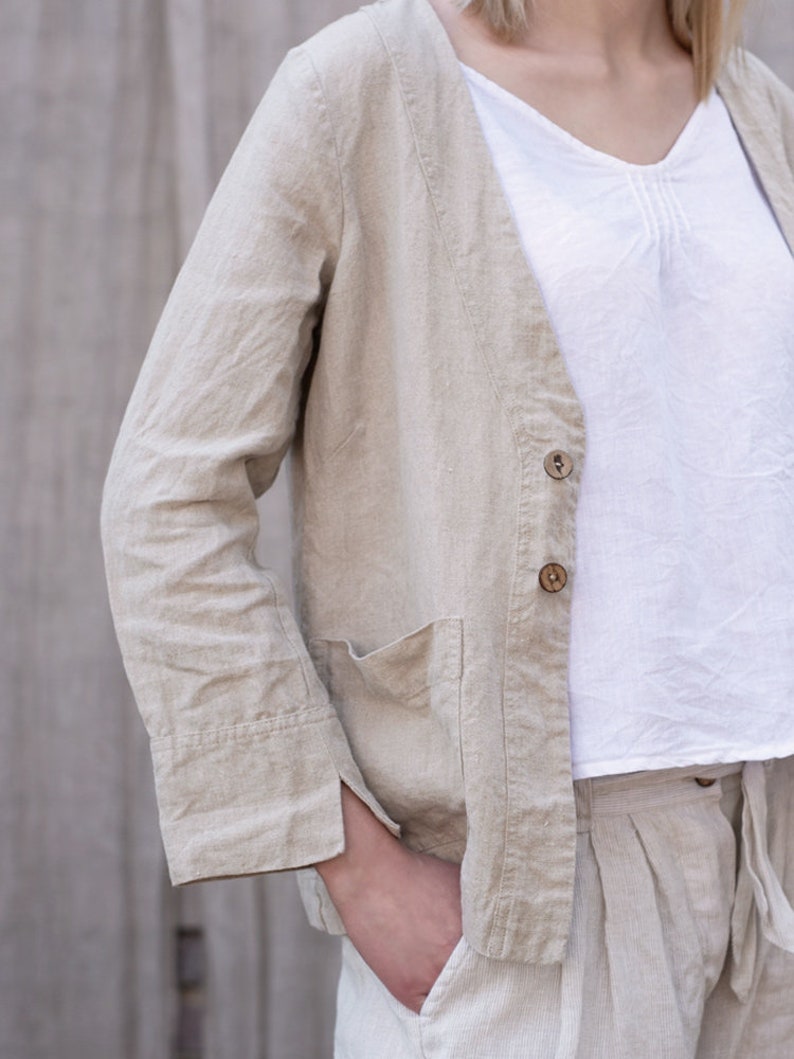 May include: A beige linen jacket with a V-neck, long sleeves, and a button closure. The jacket has a patch pocket on the left side and a single button on the front.