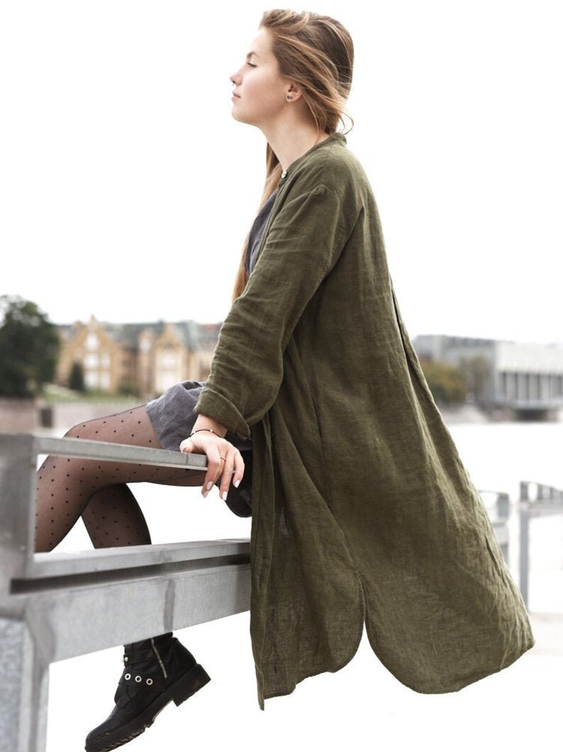 May include: A woman wearing a long, olive green linen shirt dress with a black and white polka dot pattern on her tights. She is sitting on a metal railing with one leg extended and wearing black ankle boots.