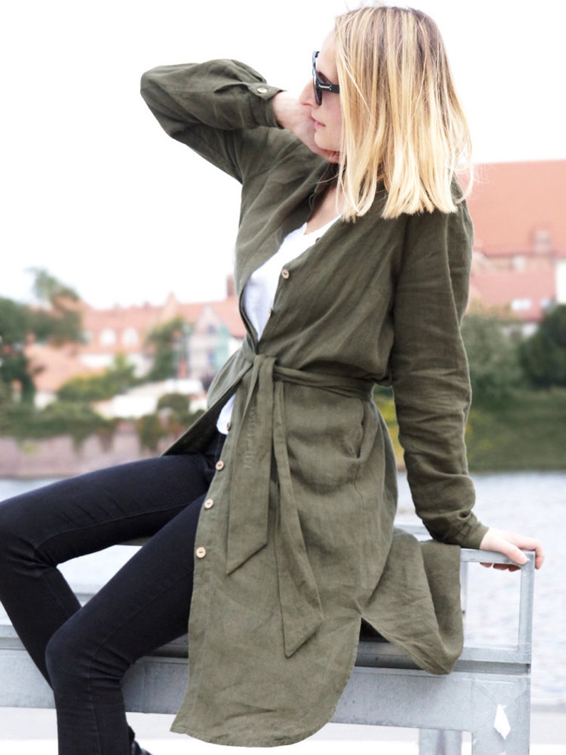 May include: A woman wearing a long, olive green linen shirt dress with a belt tied at the waist. She is wearing black jeans and sunglasses.