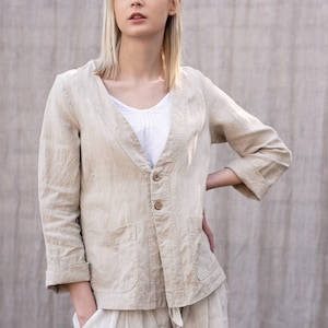 May include: A woman wearing a light beige linen jacket with a button closure and two pockets. The jacket has a V-neck and is worn over a white top. The woman is also wearing matching linen pants.