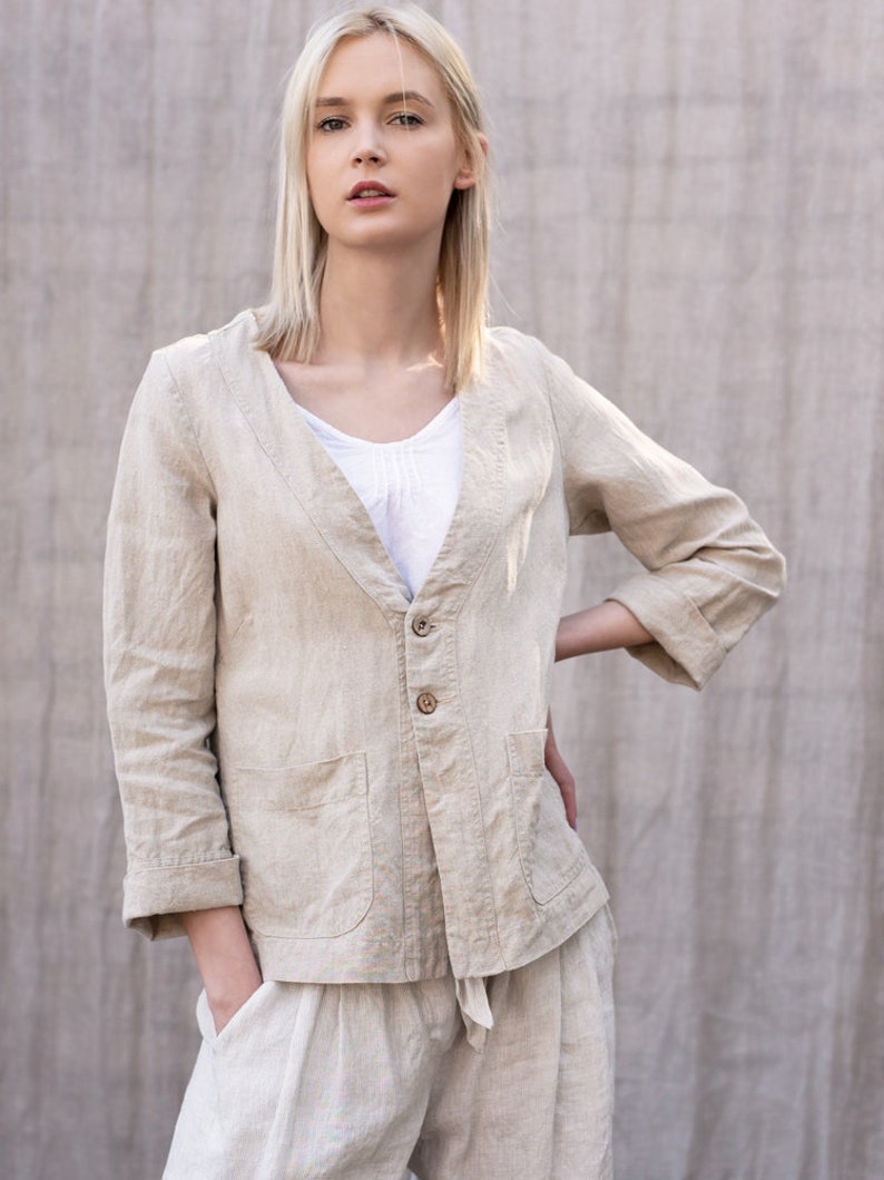 May include: A woman wearing a light beige linen jacket with a button closure and two pockets. The jacket has a V-neck and is worn over a white top. The woman is also wearing matching linen pants.