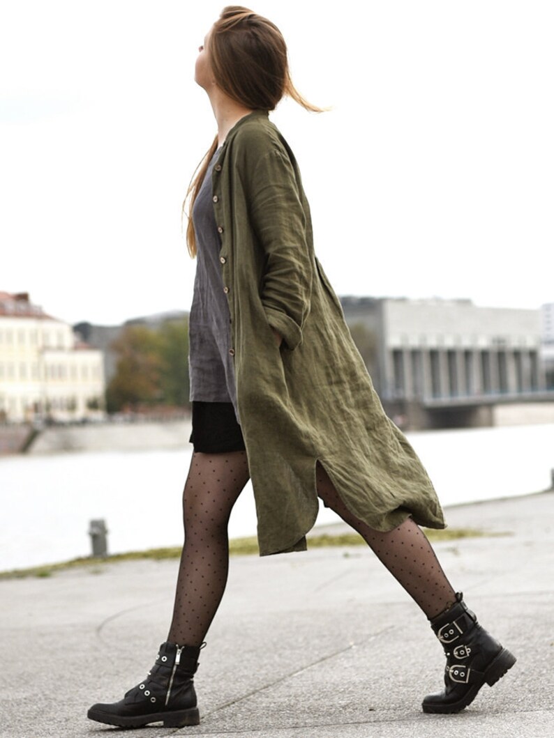 May include: A woman wearing a long, olive green linen coat over a gray shirt and black skirt. She is wearing black tights with white polka dots and black ankle boots with buckles.