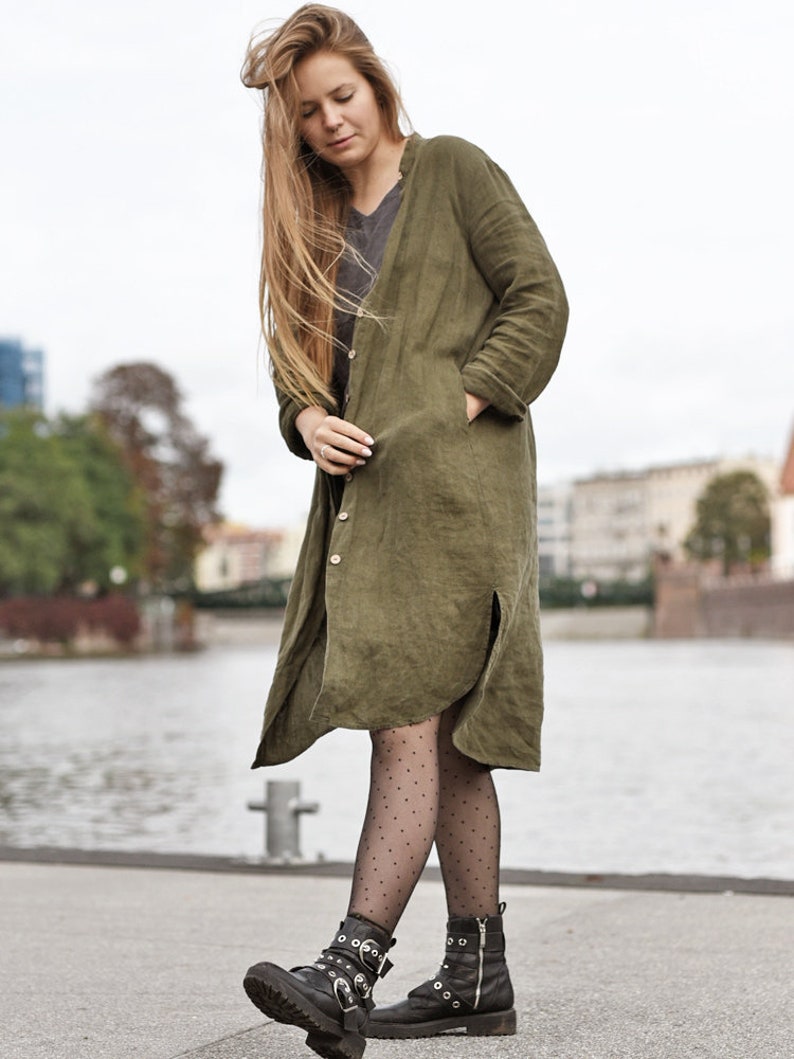 May include: A woman wearing a long, olive green linen shirt dress with a button-up front and pockets. She is wearing black tights with white polka dots and black combat boots with buckles.