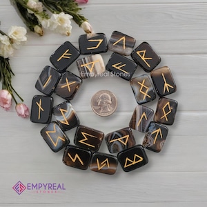 Può includere: Un set di 24 pietre di rune in agata nera con rune dorate. Le pietre sono disposte in cerchio su una superficie bianca. Le rune sono le rune dell'Elder Futhark, che sono usate nella mitologia norrena e nella magia.