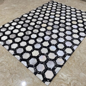 Könnte beinhalten: Ein rechteckiger Teppich mit einem geometrischen Muster aus schwarzen, weißen und grauen Sechsecken. Das Design des Teppichs weist eine wabenartige Anordnung auf, die einen modernen Look erzeugt. Geeignet für Wohnzimmer, Schlafzimmer oder Eingangsbereiche.