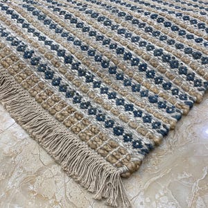 Puede incluir: Una alfombra tejida con un patrón geométrico en beige, azul y blanco. La alfombra tiene un borde con flecos y una superficie texturizada. El diseño presenta filas de colores y formas alternas, creando un textil visualmente atractivo.