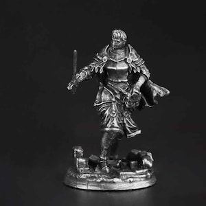 K&ouml;nnte beinhalten: Eine silberfarbene Metallminiaturfigur eines Kriegers in Plattenr&uuml;stung, die auf einem mit Tr&uuml;mmern bedeckten Sockel steht. Die Figur h&auml;lt ein Schwert in der rechten Hand und tr&auml;gt einen wehenden Umhang, mit detaillierter R&uuml;stung an Brust und Schultern.