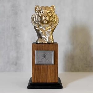 Op de afbeelding: Goudkleurige tijgerbuste op een houten voet met een zilveren plaat. De sculptuur heeft een gedetailleerde vachttextuur en staat op een donkere voet. De plaat bevat Aziatische karakters.