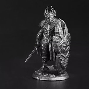 K&ouml;nnte beinhalten: Eine silberfarbene Metallfigur eines drachenartigen Kriegers, der auf einem runden Sockel steht. Die Figur tr&auml;gt eine detaillierte Plattenr&uuml;stung, hat einen geh&ouml;rnten Kopf und h&auml;lt ein langes, gerades Schwert sowie einen gro&szlig;en, strukturierten Schild.