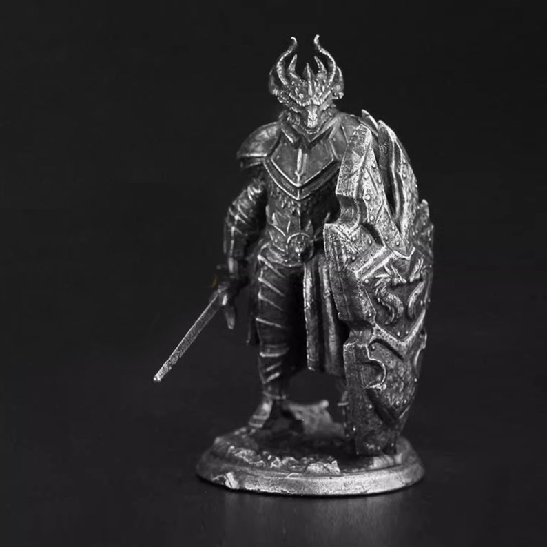 K&ouml;nnte beinhalten: Eine silberfarbene Metallfigur eines drachenartigen Kriegers, der auf einem runden Sockel steht. Die Figur tr&auml;gt eine detaillierte Plattenr&uuml;stung, hat einen geh&ouml;rnten Kopf und h&auml;lt ein langes, gerades Schwert sowie einen gro&szlig;en, strukturierten Schild.