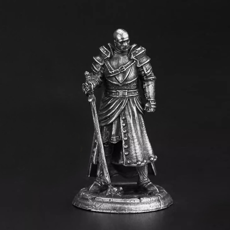 K&ouml;nnte beinhalten: Eine silberfarbene Metallminiaturfigur eines Kriegers, der auf einem runden Sockel steht. Die Figur tr&auml;gt eine detaillierte Plattenr&uuml;stung, eine lange Tunika mit Kn&ouml;pfen und h&auml;lt ein langes Schwert, dessen Spitze auf dem Boden ruht.