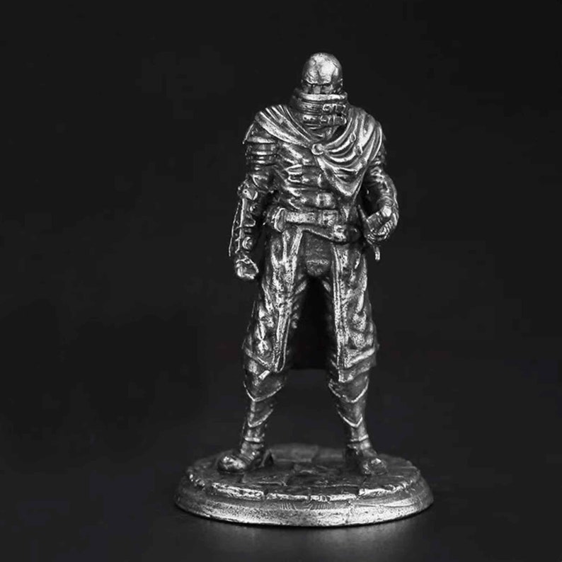 K&ouml;nnte beinhalten: Eine silberfarbene Metallfigur eines stehenden Kriegers mit texturierter R&uuml;stung, einem Umhang mit hohem Kragen und geschichteter Kleidung. Die Figur steht auf einem runden, steinstrukturierten Sockel und weist detaillierte Verzierungen auf.