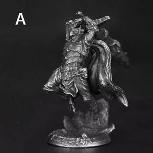 Può includere: Statuetta in scala di grigi di un guerriero con armatura dettagliata, che brandisce una spada in una posa dinamica. La figura è su una base strutturata, con un mantello fluente che aggiunge la sensazione di movimento. La lettera "A" è nell'angolo in alto a sinistra.