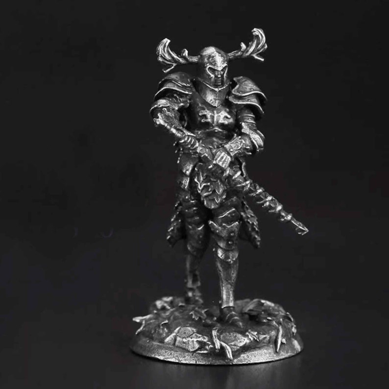 K&ouml;nnte beinhalten: Eine silberfarbene Metallminiaturfigur eines Kriegers in Plattenr&uuml;stung und einem Helm mit gro&szlig;en, verzweigten Geweihen. Die Figur steht auf einem strukturierten, felsigen Sockel und h&auml;lt ein langes, schmales Schwert, das nach unten zeigt.