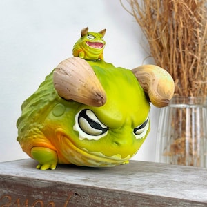Puede incluir: Una figura de monstruo verde y amarilla con cuernos y ojos grandes. Una figura de monstruo verde más pequeña con cuernos y ojos grandes se sienta en la cabeza del monstruo más grande.