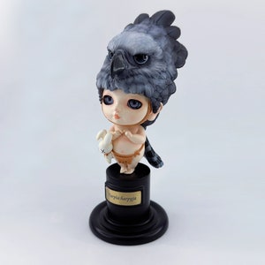 Puede incluir: Una pequeña figura detallada de un niño con cabeza de pájaro, sosteniendo un pequeño conejo blanco. La figura está de pie sobre un pedestal negro con una etiqueta dorada que dice "Harpia harpyja".