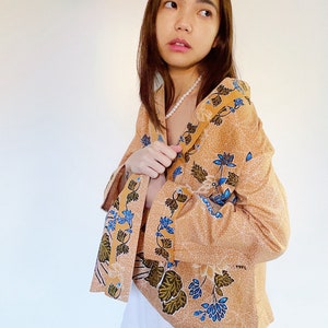 Può includere: Una donna indossa una blusa marrone chiaro con un motivo floreale in tonalità blu e verdi. La blusa ha maniche lunghe con polsini larghi e una vestibilità rilassata. Indossa una collana di perle bianche.