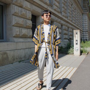 Può includere: Una persona indossa una giacca in stile kimono fantasia con strisce gialle, blu navy e grigie, abbinata a una maglietta bianca e pantaloni grigio chiaro. L'outfit è completato da sandali neri. Lo sfondo presenta un edificio in pietra e un vialetto lastricato.