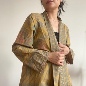Op de afbeelding: Een geel, groen en bruin gepatroneerd jasje in kimono-stijl met een lange, open voorkant en lange mouwen. Het jasje heeft een bloemen- en geometrisch ontwerp.