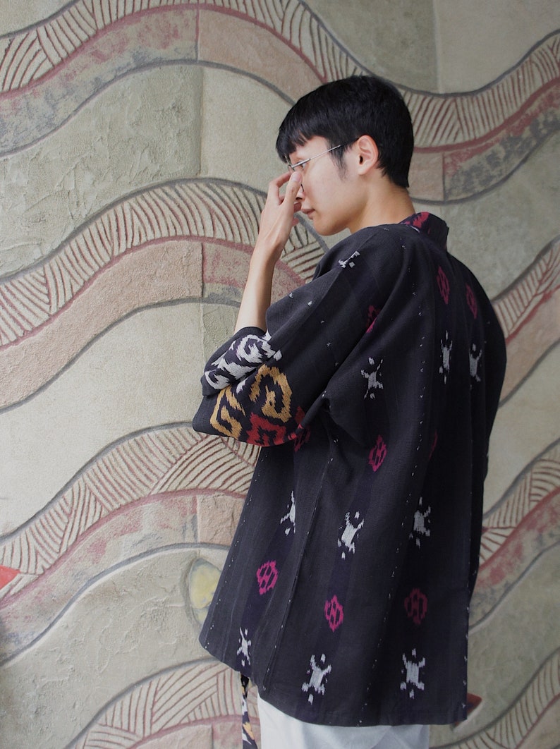 May include: A dark blue kimono-style jacket with a patterned design in white, pink, and yellow. The sleeves have a contrasting pattern. The jacket is worn by a person.