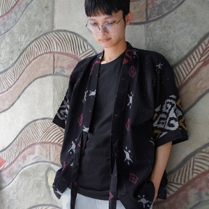 May include: A black kimono-style jacket with a patterned design in red, white, and gray. The jacket is worn over a black t-shirt and paired with light gray pants. The outfit is casual and stylish.