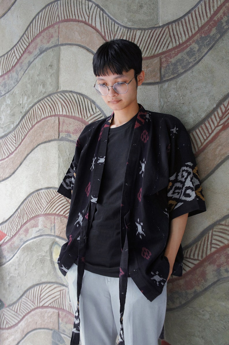 May include: A black kimono-style jacket with a patterned design in red, white, and gray. The jacket is worn over a black t-shirt and paired with light gray pants. The outfit is casual and stylish.