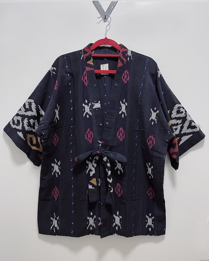 May include: A dark blue kimono-style jacket with a patterned design. The jacket features a V-neck, short sleeves, and a tie closure. The pattern includes white, pink, and yellow accents. The jacket is hanging on a red hanger.