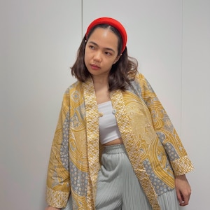 Può includere: Una donna indossa una giacca in stile kimono con motivo giallo e grigio con un top bianco sotto. Indossa anche una gonna plissettata blu chiaro e una fascia per capelli rossa.