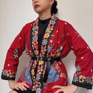 Op de afbeelding: Een rode kimono met een zwarte bies en een bloemenpatroon. De kimono is gegespt met een zwarte leren riem. De kimono heeft een pauwenmotief aan de voorkant.