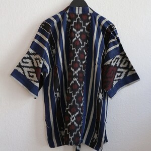 K&ouml;nnte beinhalten: Blau-wei&szlig; gemusterte Kimono-Jacke mit kurzen &Auml;rmeln, die an einer wei&szlig;en Wand h&auml;ngt. Die Jacke hat vertikale Streifen und ein zentrales Design mit roten und wei&szlig;en geometrischen Formen. Ein Bindeg&uuml;rtel h&auml;ngt von unten.