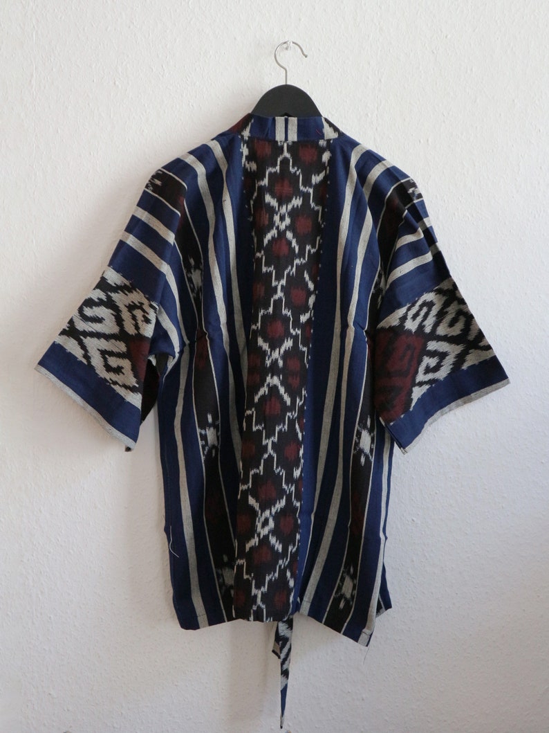 K&ouml;nnte beinhalten: Blau-wei&szlig; gemusterte Kimono-Jacke mit kurzen &Auml;rmeln, die an einer wei&szlig;en Wand h&auml;ngt. Die Jacke hat vertikale Streifen und ein zentrales Design mit roten und wei&szlig;en geometrischen Formen. Ein Bindeg&uuml;rtel h&auml;ngt von unten.