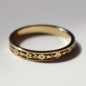 May include: A gold ring with a floral design. The band of the ring is decorated with a repeating pattern of small flowers and leaves. The ring is shown against a white background, with light reflecting off the polished surface.