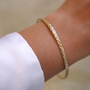 Peut inclure: Un bracelet jonc fin de couleur dorée présentant une texture torsadée en forme de corde continue. Le bracelet est de forme circulaire et possède une finition métallique polie et réfléchissante qui capte la lumière.