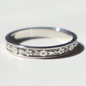 May include: A silver ring with a floral pattern. The ring has a band of white flowers and leaves around the edge. The ring is set against a white background.
