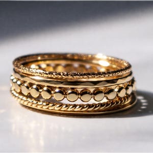 May include: A stack of gold rings with various textures. One ring has a rope design, another has small circular accents, and others have a hammered or textured surface. The rings are displayed on a white surface.