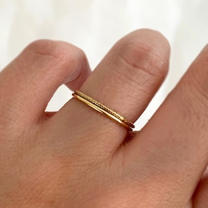 May include: Two gold rings on a finger. The top ring has a textured design, while the bottom ring is smooth.
