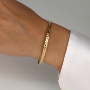 May include: A gold bangle bracelet with an engraved floral design. The bracelet is worn on a wrist, with a white shirt sleeve in the background. The bangle is a simple, elegant piece of jewellery.