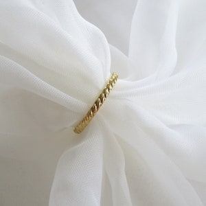 May include: A gold-colored ring with a braided design. The ring is on a white fabric background.