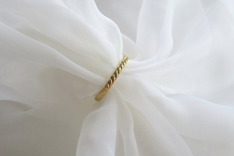 May include: A gold-colored ring with a braided design. The ring is on a white fabric background.