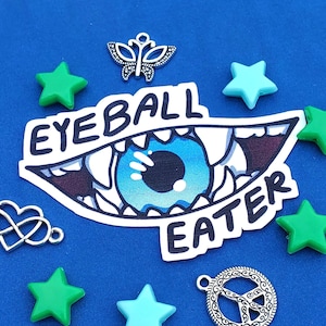 May include: A blue and white eye with teeth and the words "Eyeball Eater" on a blue background. There are several green and blue stars, a silver butterfly charm, a silver infinity heart charm, and a silver peace sign charm.