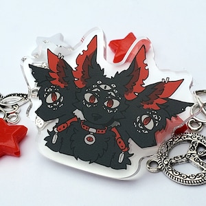 May include: A clear acrylic charm featuring a black and red three-headed creature with multiple eyes. The charm is attached to a silver peace sign charm.