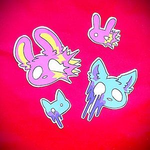 May include: Four stickers featuring stylized animal heads. Two pink rabbit-like heads with yellow accents and white eyes, and two blue cat-like heads with purple dripping effects and white eyes, all arranged against a solid bright red background.