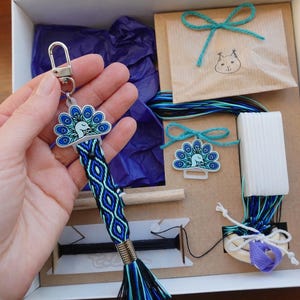 Könnte beinhalten: Ein Pfau-Schlüsselanhänger mit blauem und weißem Design, gehalten in einer Hand, ist der Mittelpunkt dieses Bildes. Der Schlüsselanhänger ist an einem blau-schwarzen gewebten Armband befestigt. Das Bild zeigt auch ein Bastelset mit verschiedenen Materialien.